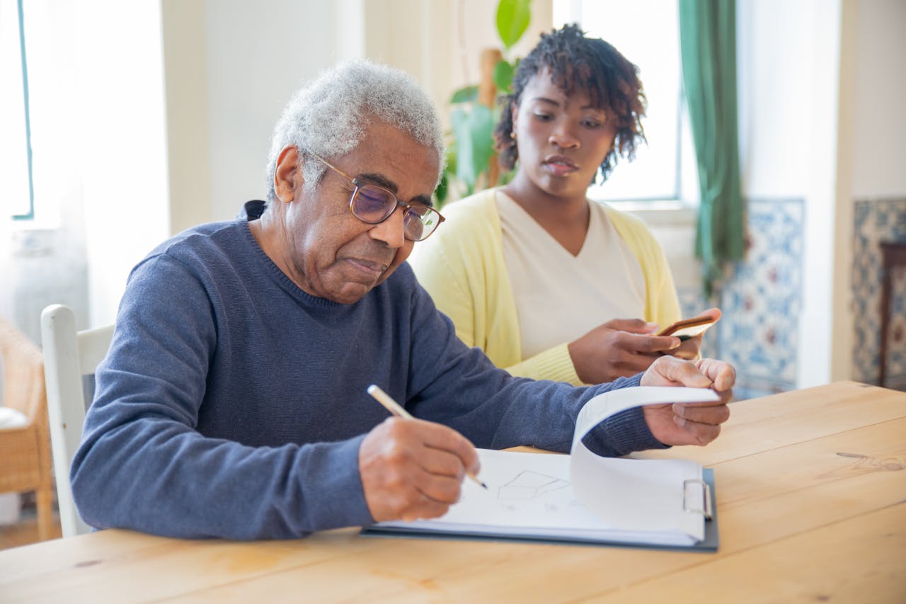 hero-gallery-02 A senior man writing on papers with a caregiver nearby indoors in a supportive setting.